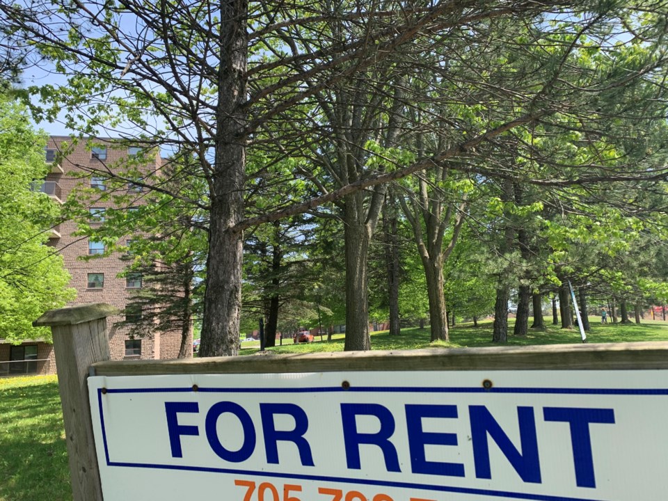 A 'For Rent' sign in front of a wooded area and residential apartment building
